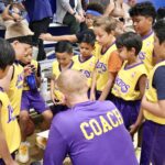 Youth basketball team huddles around coach in purple and gold jerseys.