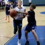 Young boys playing basketball, one driving to the hoop while the other defends.