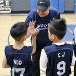 Coach high-fives two young basketball players wearing jerseys in a gym.