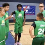 Youth basketball team in green jerseys listening to coach indoors.