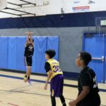 Two boys playing basketball in a gymnasium, one shooting a jump shot.