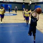 Young children playing basketball in a gymnasium, with one child attempting a layup.