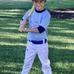Young boy in baseball uniform holding a bat and smiling outdoors.