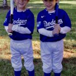 Two young boys in Dodgers baseball uniforms holding bats and smiling outdoors.