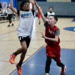Two young boys playing basketball indoors, one attempting a shot while the other defends.