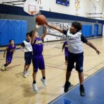 Children playing basketball in a gym, one girl defending a boy shooting.