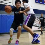 Two boys playing basketball indoors, one dribbling past the other.