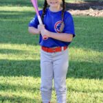 Young girl in baseball uniform holding a bat on grass.