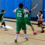 Youth basketball players in green and black uniforms playing on an indoor court.