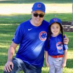 A man and child smiling in Chicago Cubs uniforms outdoors.
