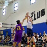 A basketball player in white jumps to shoot over a defender in purple.