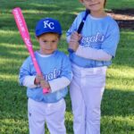Two young boys in Kansas City Royals baseball uniforms, one holding a pink bat.