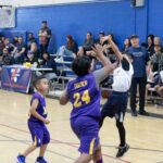 Kids playing basketball in a gymnasium with spectators watching.