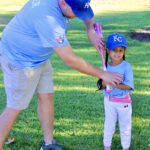 A man helps a young girl learn to swing a baseball bat in a sunny park.