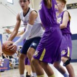 Two basketball players in purple jerseys defend a player in a white jersey holding the ball.