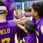 Young basketball players in purple Lakers jerseys join hands in a huddle.