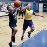 Two kids playing basketball in a gym, one attempting a layup.