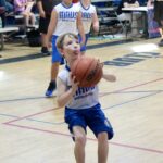 Young boy in blue and white jersey shooting a basketball in a gym.