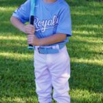 A young baseball player in a blue Royals jersey holding a bat on a grassy field.