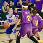 Youth basketball player in blue dribbles past defenders in purple jerseys.