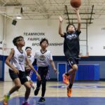 Kids playing basketball indoors, one jumping to shoot.