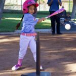 Young girl playing tee-ball, hitting the ball with a pink bat.