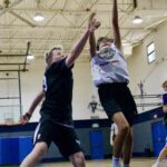 Two boys playing basketball indoors, one jumping to shoot while the other defends.