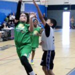 Young basketball player in green leaps for a shot against defenders.