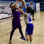 Two kids playing basketball indoors, one in a purple Lakers jersey.