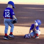 Two kids playing baseball, one standing and one sitting on the ground.