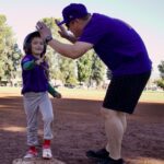 A man helps a young girl with baseball batting stance outdoors.