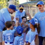 Kids in blue baseball uniforms gathered around coaches on a sunny field.