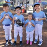 Four children in baseball uniforms stand on a field holding trophies.