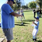 A man helps a young boy with baseball practice in a park.