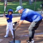 Young boy batting in a tee-ball game with adult coach.