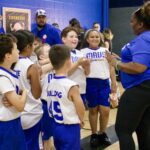 Cheerful kids in sports uniforms gather around their coach indoors.